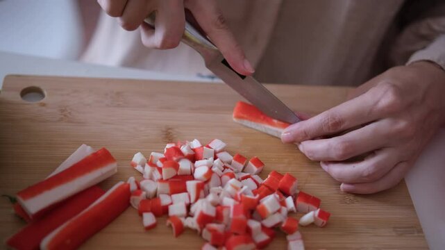 Female hands cutting red crab sticks or surimi with knife on a wooden cutting board close up. Cooking at home