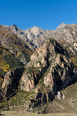 Kabardino-Balkaria. El-Tyubu. Rocky mountains with trees growing on their slopes glow in morning sun. Close-up. Chegem Gorge. Unique rocky ridge with steep cliffs against blue autumn sky.