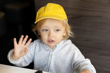 a 3-year-old boy dressed in a yellow cap is waving his hand