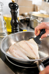 A chef skillfully flips fish fillets in a frying pan, showcasing the process of cooking in a professional kitchen. The scene captures culinary expertise and fresh ingredients.