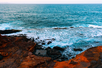 Rocky coastline with waves crashing against the shore. Water appears to be mix of blue and white due to foam created by waves. Rugged rocks and dynamic ocean waves makes beauty of coastal landscapes