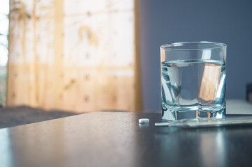 in the room, on the bedside table, there is a glass of yellow medicine next to a thermometer and a vitamin C tablet. High quality photo