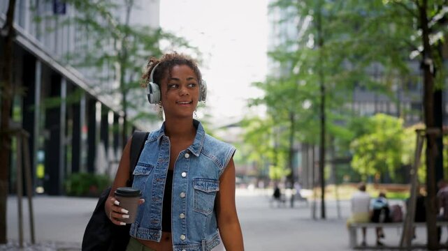 Young woman walking through the city with backpack on back. University student is heading to class, holding a coffee in a travel cup.