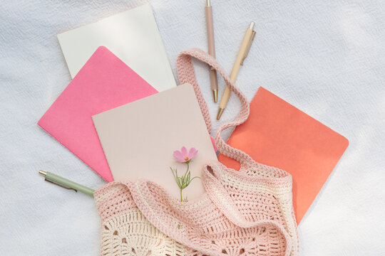 Overhead view of assorted notebooks, pens and a pink flower in a pink knitted bag on white fabric