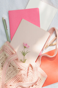 Overhead view of assorted notebooks, pens and a pink flower in a pink knitted bag on white fabric