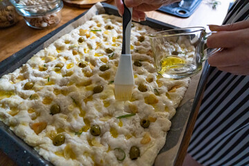 Woman greasing raw dough with olive oil. Uncooked focaccia alle olive - foccacia with green olives,...