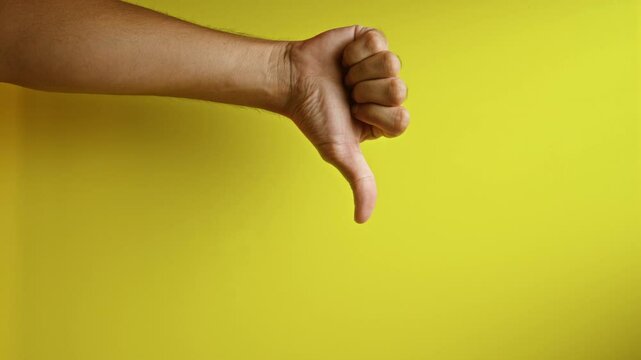 Man showing thumbs down gesture on yellow background with hand clearly visible portraying disapproval and negative sentiment