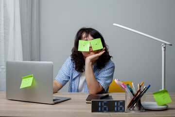 A tired woman with curly hair in a striped shirt sits at a cluttered desk with sticky notes covering her eyes. She rests her head on her hand, surrounded by a laptop, clock, lamp, and stationery.