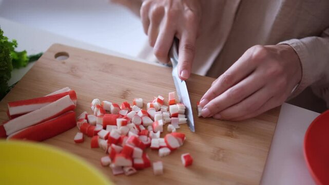 Female hands cutting red crab sticks or surimi with knife on a wooden cutting board close up. Cooking at home
