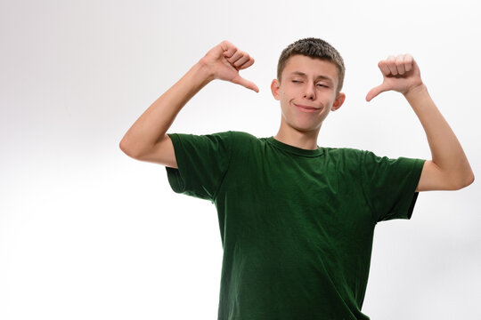 A young man in a green t-shirt stands against a white background, using his hands to gesture towards himself in a self-deprecating or unimpressed manner.