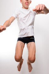 A low-angle shot of a teenage boy mid-jump against a light gray background. He's wearing a white t-shirt and dark shorts. The image captures his dynamic movement and youthful energy.