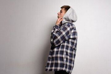 A young person wearing a blue and white plaid shirt over a gray hoodie. He's adjusting the hood, standing against a plain white wall. The overall mood is casual and relaxed.