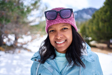 Cheerful portrait of a young African American woman standing in a snowy forest enjoying the sunny day outdoors, looking at camera smiling. Copy space.