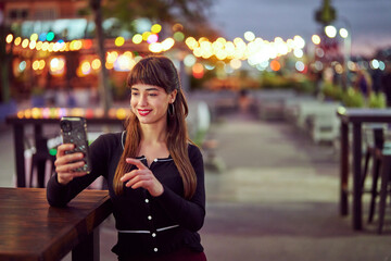 Smiling caucasian woman using mobile phone while sitting at a table in a street cafe at night in Puerto Madero, Buenos Aires 