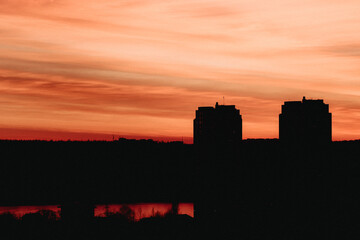 Silhouette of two tall buildings against a dramatic orange-red sunset sky with streaks of clouds. The lower part of the image is dark, with reflections of the sunset visible on a body of water.