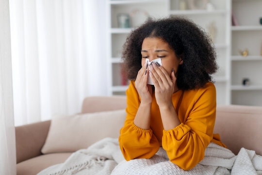 Unwell young african american woman blowing her nose into tissue at home, sick african american female wrapped in blanket sitting , suffering symptoms of cold and flu, copy space. High quality photo