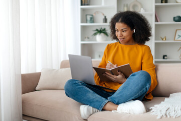 Portrait of smiling woman sitting on sofa, using laptop and writing in notebook, taking notes,...