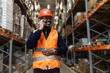 Warehouse worker with safety vest and helmet talking on mobile phone in logistics distribution...