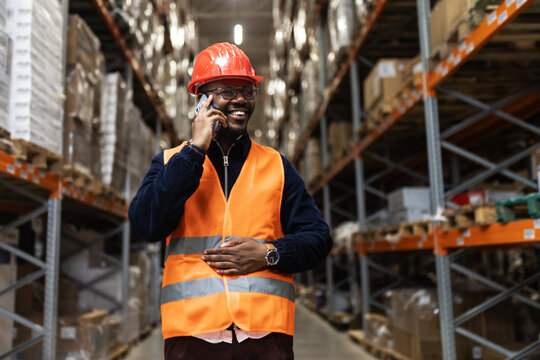 Warehouse worker with safety vest and helmet talking on mobile phone in logistics distribution center. Confident professional managing inventory and communication 