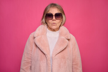 An elderly woman confidently poses against a bright pink background. She is wearing oversized sunglasses and a fluffy light pink coat, showcasing her stylish flair and attitude