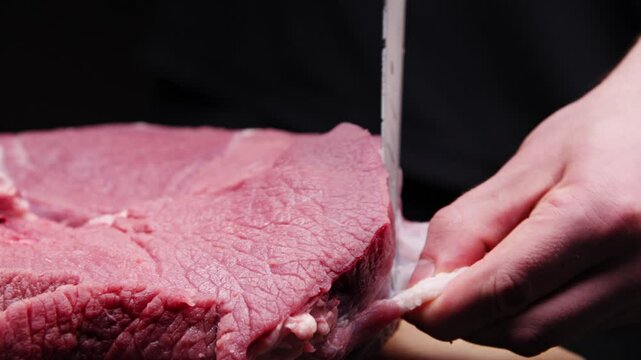 A butcher removes the fascia from a beef steak with a large knife in a dark room