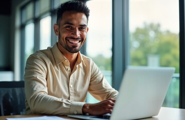 Fototapeta premium Happy Hispanic businessman sits at desk in office, smiles at camera, working on laptop. Successful pro, likely manager, looks satisfied. Modern office setting. Image successful employee. Focus on