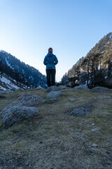 A female wearing a blue down jacket and colorful woolen cap stands facing a serene snow-covered mountain valley. The rocky terrain, scattered stones, pine trees, a clear blue sky and peaceful nature.