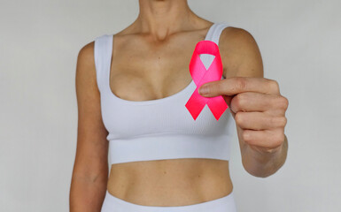 Woman wearing a white top holds a pink ribbon highlighting breast cancer awareness in a neutral...