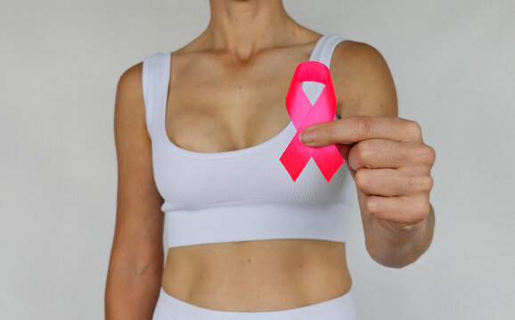 Woman wearing a white top holds a pink ribbon highlighting breast cancer awareness in a neutral setting