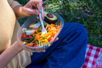 A person enjoying a fresh salad with avocado while sitting on a picnic blanket in a sunny park setting