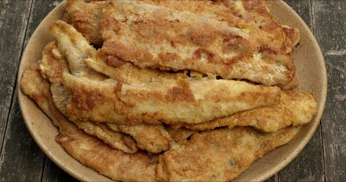 A pile of fried pike-perch fillets coated in flour and egg on a plate. Top view, table spin.
