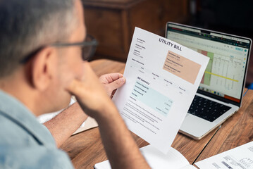 Man examining utility bill at home office with laptop and documents on wooden table. Concept of household finance, bill management, and monthly budgeting 