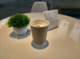 In some cafe, there is a paper cup with freshly brewed coffee on the table, a napkin holder, and an ornamental plant, a symbolic and simple still life from a coffee shop