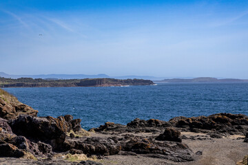 A breathtaking seascape showcasing a vast expanse of the blue ocean bordered by rocky outcrops in the foreground. The distant coastline features layered cliffs and rolling hills under a clear blue sky