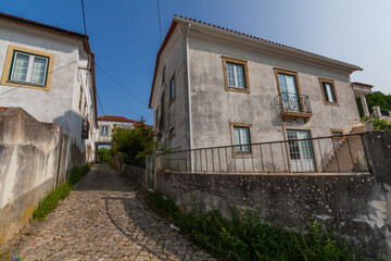 A narrow cobblestone street winds between traditional white buildings with colorful window frames under a clear blue sky