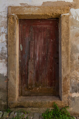 Fototapeta premium A weathered, dark red wooden door with a stone frame is set into a crumbling wall, with weeds growing at the base