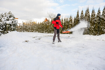 Individual in red jacket shovels snow in a snowy area with tall evergreens, bare trees, rooftops,...