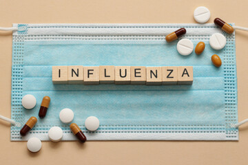 Influenza spelled with wooden cubes on a surgical mask with scattered pills around.