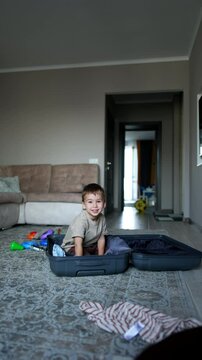 Happy toddler boy gets into the big suitcase. Kid stands up and picks some of his clothes scattered around the room. Vertical video.