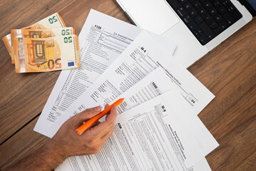 Fototapeta premium Close-up of a man filling out tax forms with euro banknotes and a laptop on a wooden table. Tax preparation and financial management concept. Ideal for tax season and accounting visuals 