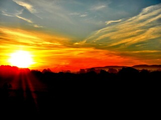 Red, orange, yellow and blue sky with clouds and sun during sunrise over dark misty horizon with mountains and trees - abstract morning autumn view. Topics: air space, weather, meteorology, wallpaper