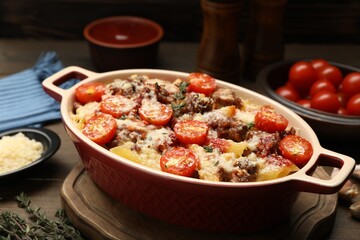 Delicious pasta casserole with cheese, tomatoes, minced meat and thyme in baking dish on wooden table, closeup
