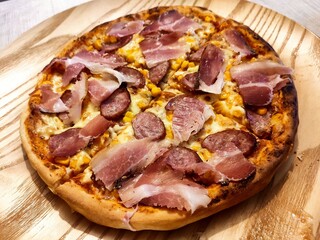 A freshly baked pizza sits on a wooden platter, generously topped with slices of sausage, bacon, and other golden-brown ingredients.