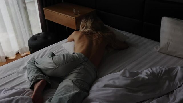 Sexy shirtless blonde in jeans with long wavy hair lying down on a pillow on a bedroom bed