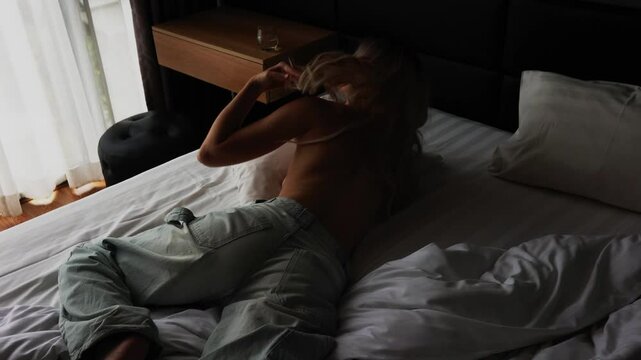Sexy blonde with long wavy hair is comfortable on pillow lying on bed in jeans