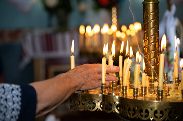 Hand of an elderly woman. Flame of lit candles. Many yellow wax candles on a candlestick. Holy...