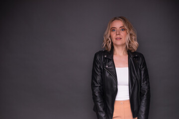 A confident blonde woman wearing a black leather jacket, white top, and beige pants poses against a...