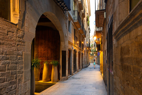 Fototapeta A young woman walks alone down a narrow alley of illuminated shops at dusk in the central Gothic Quarter of Barcelona, Spain.