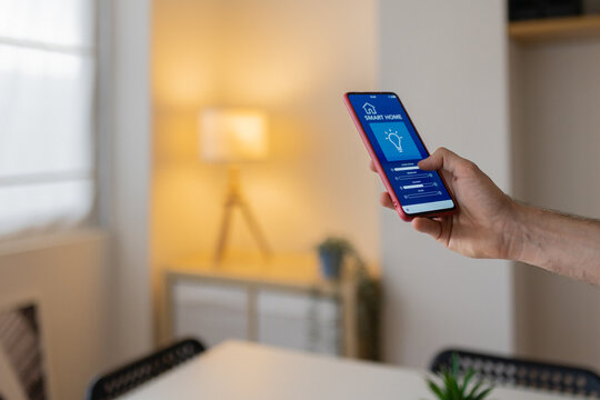 Hand holding smartphone and adjusting light intensity with smart home application in a modern apartment. IOT concept
