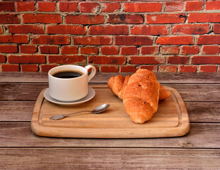 Cup of hot black coffee dessert spoon and two fresh croissants on a wooden tray on the table.